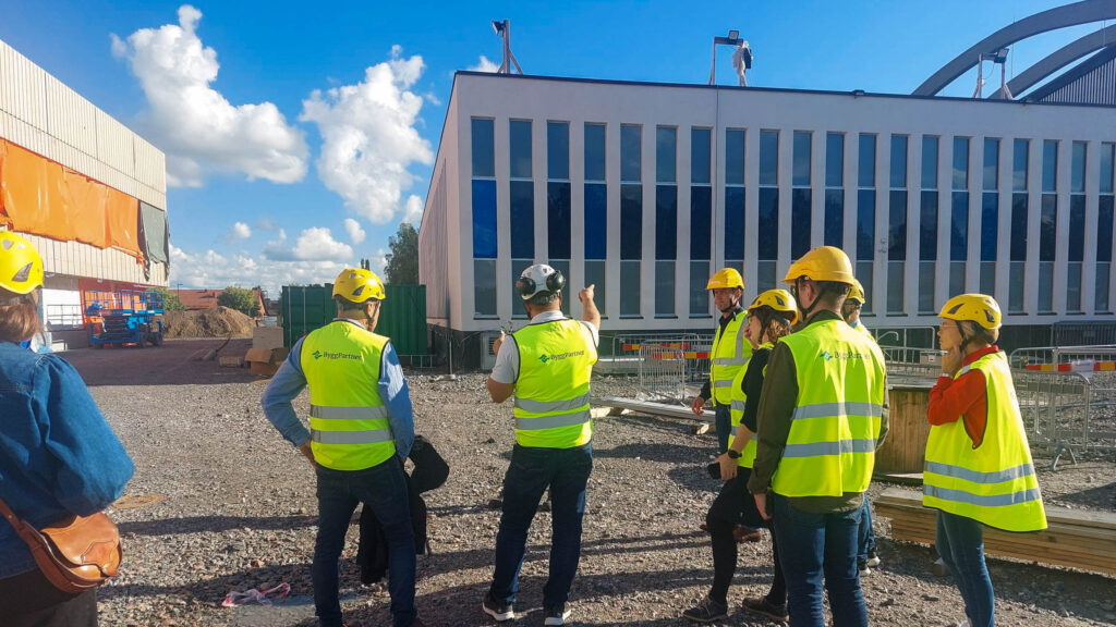 Fem personer står med skyddshjälp och reflexvästar på sig framför nybyggnationen av nya Maserhallen i Borlänge.